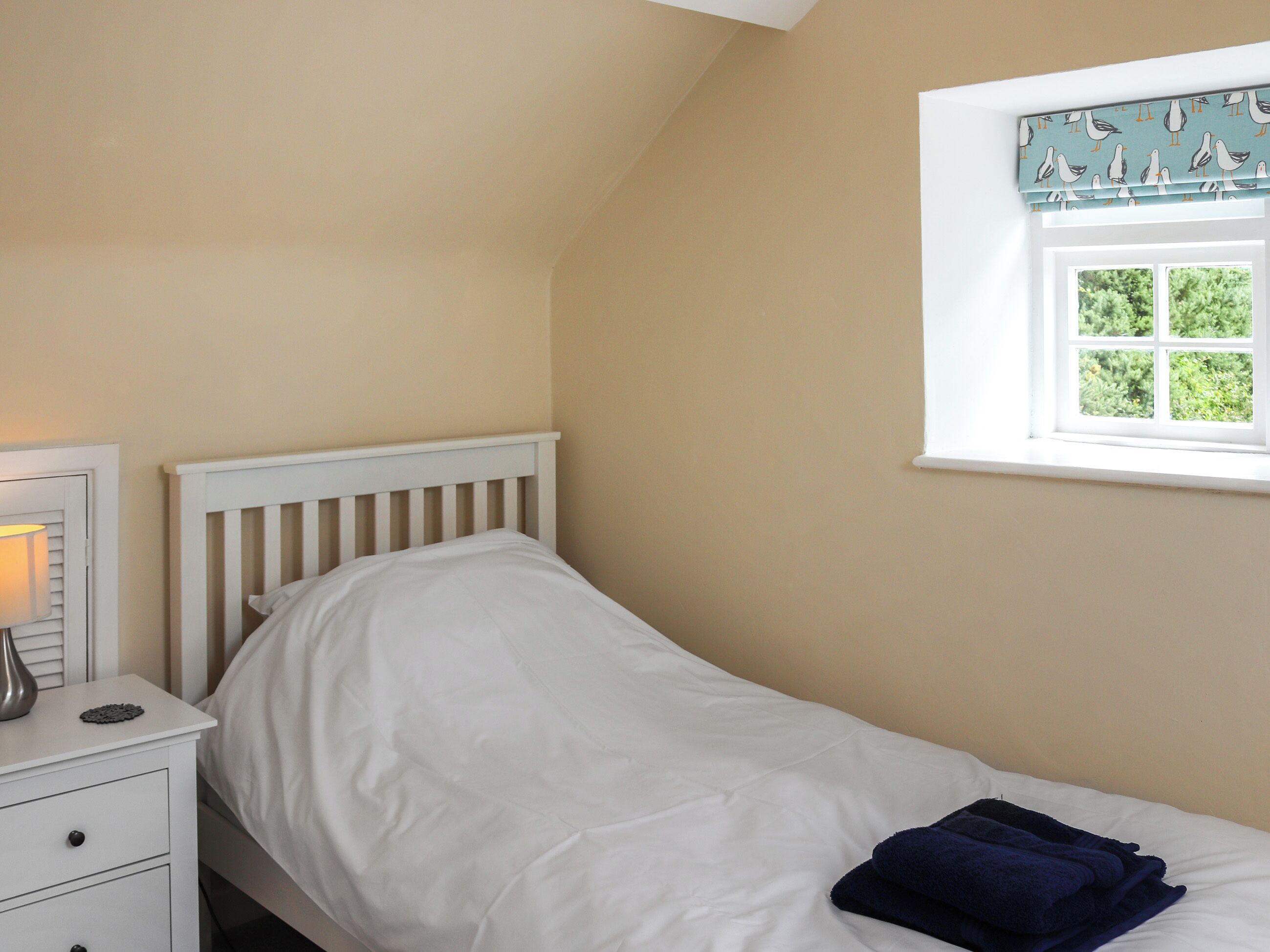 Une chambre avec un lit simple, une table de chevet avec une lampe et une fenêtre agrémentée d’un lambrequin orné de motifs de mouettes.