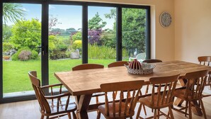 A dining area with a large table and chairs, a clock on the wall, and a view of a garden through large sliding glass doors. - 4 bedroom accommodation in Tudweiliog (Tudweiliog)