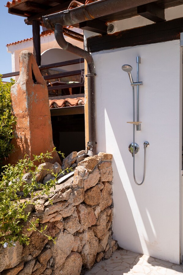 A stone wall with a metal showerhead attached to a white wall, a rustic chimney, and a tiled roof structure.