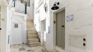 A narrow alleyway with a staircase leading to a door, a potted plant on the balcony, and a sign above the door.