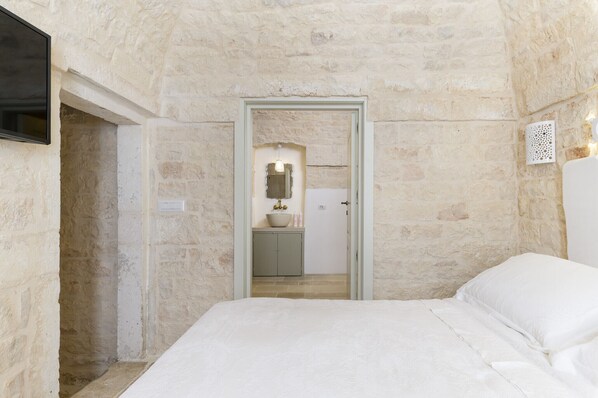 A bedroom with a stone wall, a bed, a TV, and a bathroom visible through an open door.