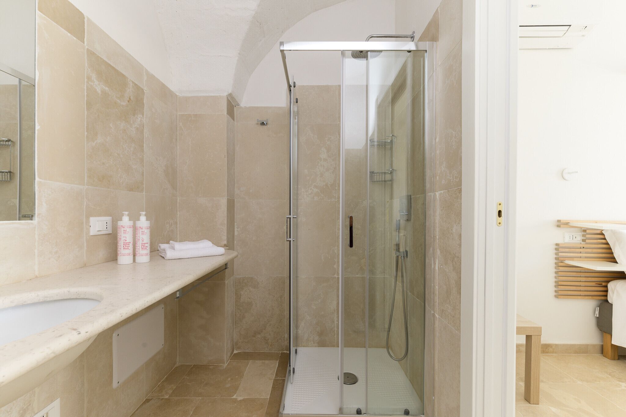 Une salle de bain moderne avec une douche à parois de verre, des carreaux de couleur beige, un lavabo blanc et un banc en bois.