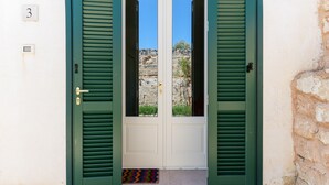 Une porte à deux battants avec des volets verts, une plaque portant le numéro « 3 » au-dessus, et un mur de pierre sur la droite.