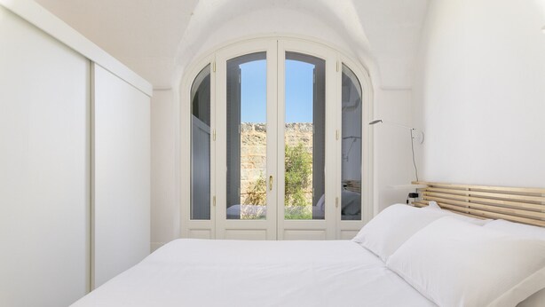 Une chambre à coucher avec un lit, des murs blancs, une tête de lit en bois et une vue sur un espace extérieur, accessible par une porte vitrée.
