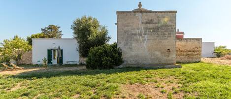 Un edificio rústico con un pequeño jardín, una estructura blanca con puerta verde y una torre de piedra.