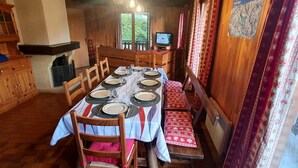 Un coin repas avec une longue table dressée pour le repas, des chaises en bois, un téléviseur et une fenêtre donnant sur un paysage verdoyant.