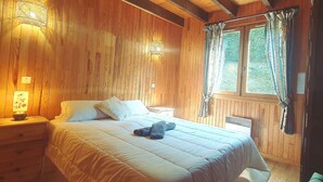 Une chambre à coucher aux murs lambrissés, avec un lit, deux lampes fixées au mur, une fenêtre avec des rideaux et une table de chevet avec une lampe.