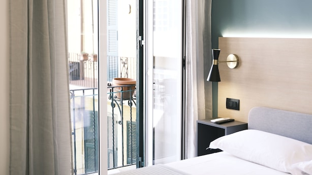 Ein Hotelzimmer mit einem Bett, einem Nachttisch, einer Wandlampe und Blick auf einen Balkon mit Geländer und einer Pflanze.