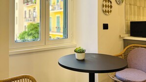 Ein Zimmer mit großem Fenster, einem runden Tisch mit Topfpflanze, zwei Korbstühlen und einem Fernseher auf einem Regal.