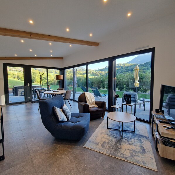 A modern living room with a sofa, armchair, and coffee table, large windows offering a view of a green landscape.