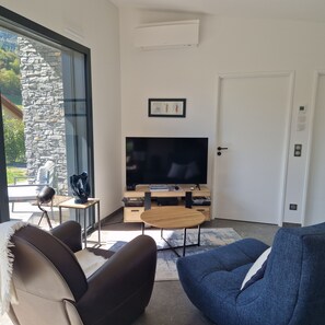 A living room with a television, a stone wall, a door, and a view of greenery outside.