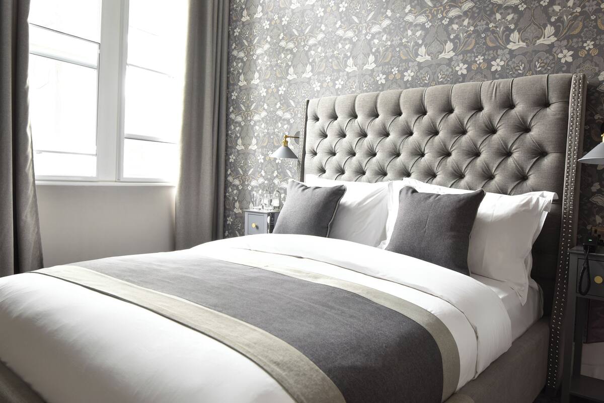 A neatly made bed with a tufted headboard, a bedside table with a lamp, and a floral wallpaper pattern.
