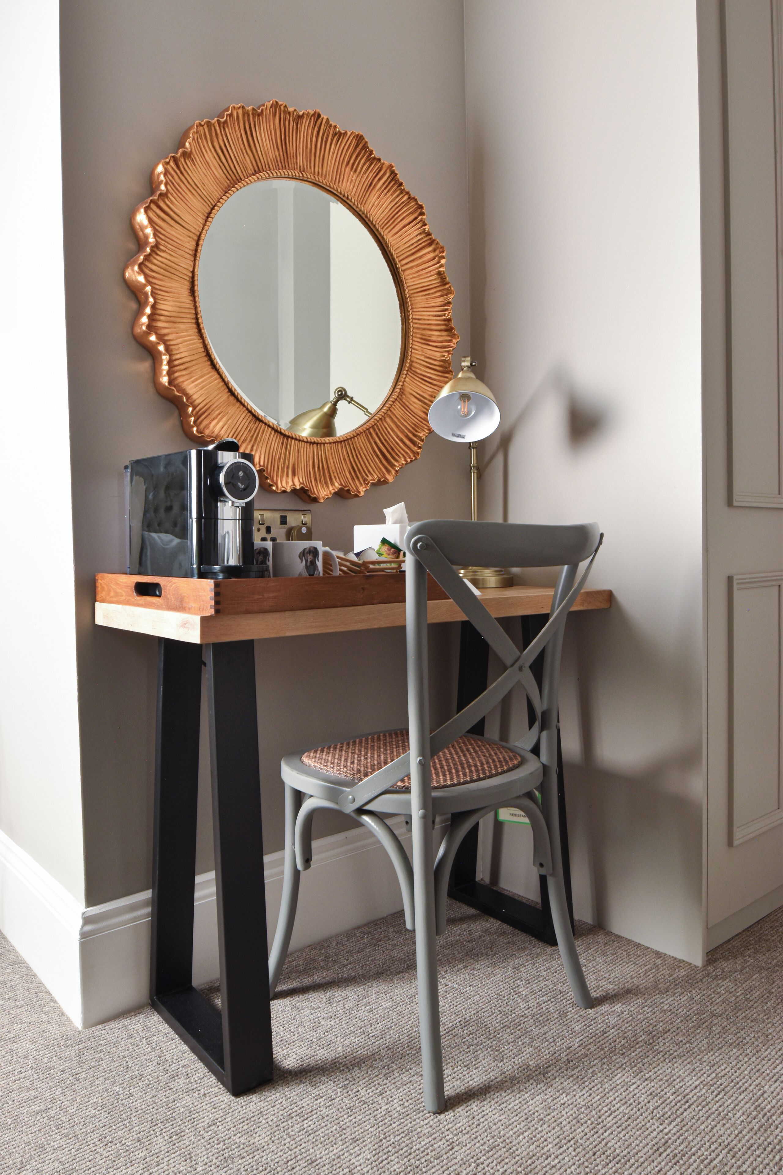 A compact workspace with a wooden desk, a grey chair, a large mirror with a decorative frame, a lamp, and a coffee maker.