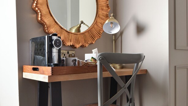 A compact workspace with a wooden desk, a grey chair, a large mirror with a decorative frame, a lamp, and a coffee maker. - Yorkshire Grey Pub & Rooms (London)