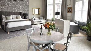 A hotel room with a large bed, a sofa, a dining table with chairs, a chandelier, a large window with curtains, and a bathtub. - Yorkshire Grey Pub & Rooms (London)