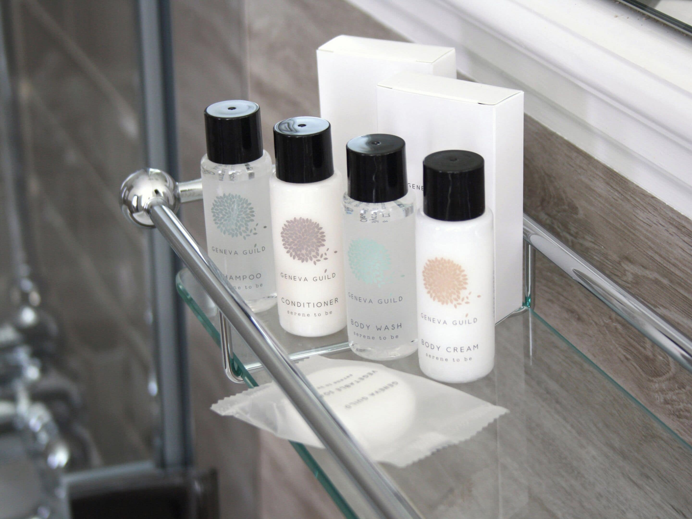 A bathroom shelf with four bottles of Geneva Guild body wash, conditioner, and shampoo, and a white box.
