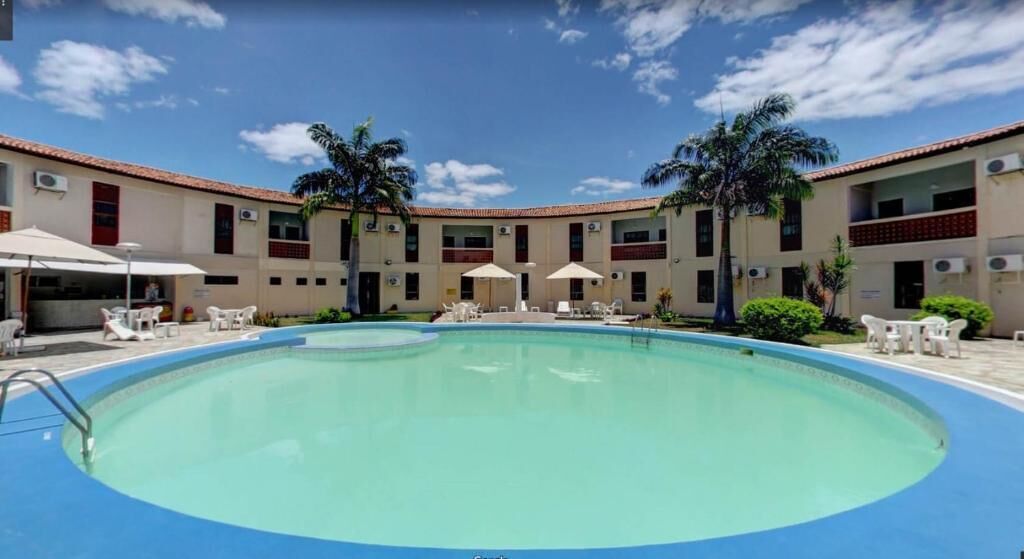 A swimming pool surrounded by lounge chairs and umbrellas, with a building featuring multiple balconies and air conditioning units in the background.