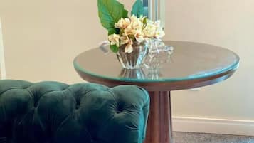Una mesa redonda de cristal con una lámpara azul oscuro, un jarrón con flores blancas y un sofá verde con capitoné.