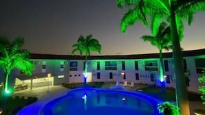 Un espace piscine nocturne avec une piscine circulaire, des palmiers et un bâtiment doté de nombreuses fenêtres.