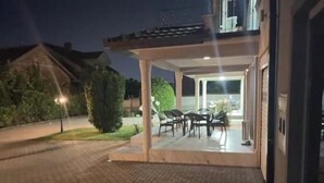 Une terrasse extérieure couverte, avec une table et des chaises, une pelouse bien entretenue et un bâtiment en arrière-plan.