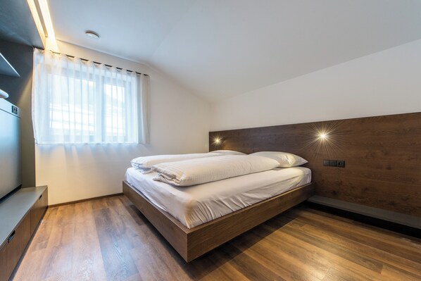 A modern bedroom with a wooden headboard, a bed with white linens, a bench, and a window with sheer curtains.