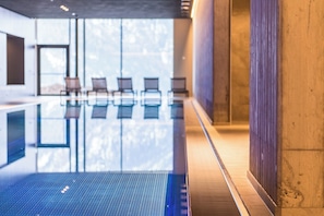 An indoor swimming pool with clear glass walls, a wooden floor, and a seating area with chairs.