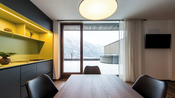 Ein moderner Essbereich mit einem Holztisch und Stühlen, einer eingebauten Regalwand mit Pflanzen, einem großen Fenster mit Bergblick und einem an der Wand befestigten Flachbildfernseher.