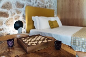 Ein Holztisch mit Schachbrett und zwei dekorativen Tassen, ein Bett mit gelbem Kopfteil und weißer Bettwäsche sowie eine Steinsäule im Hintergrund.