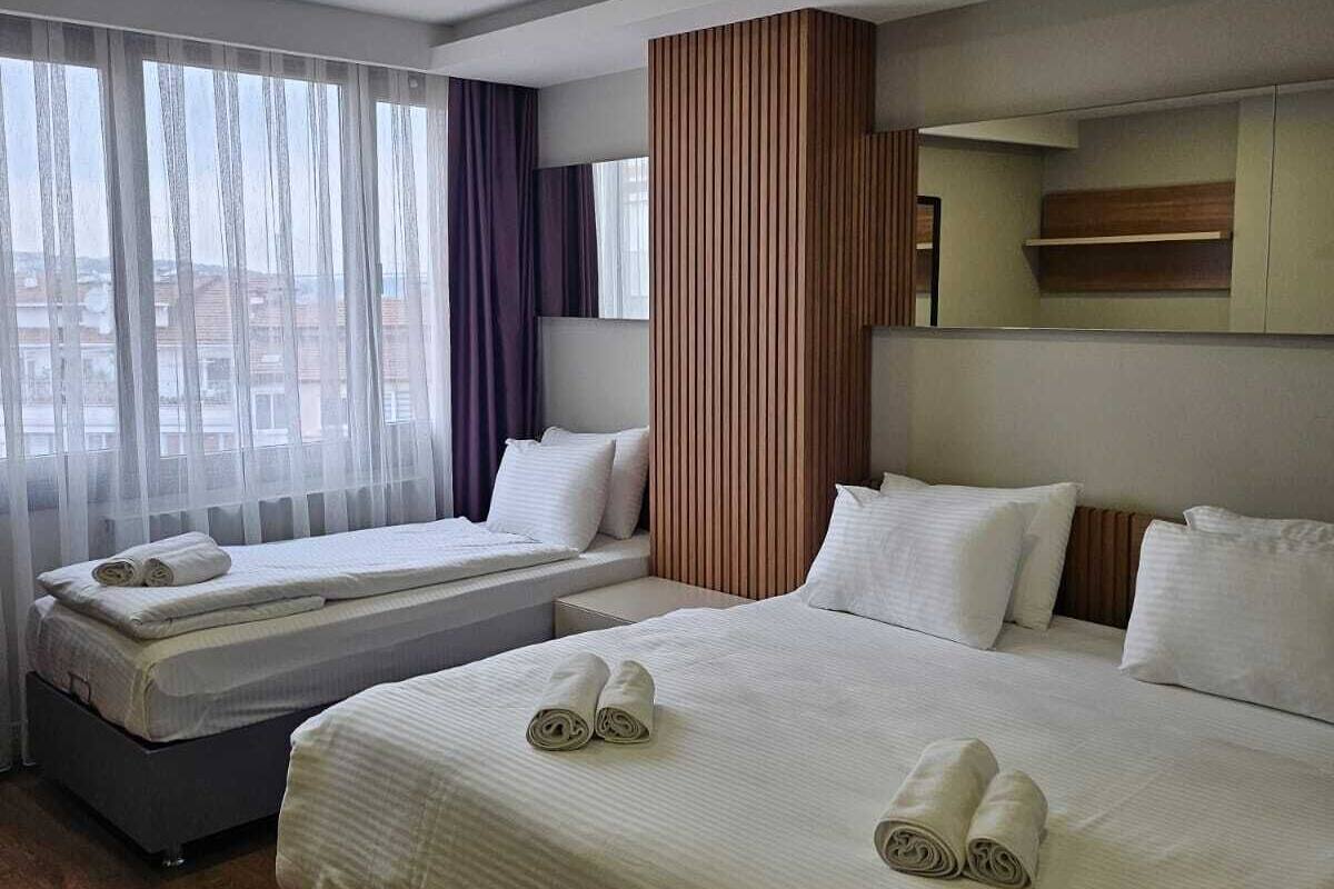 Une chambre d’hôtel avec deux lits, une grande fenêtre avec des rideaux légers et une vue sur les bâtiments environnants.