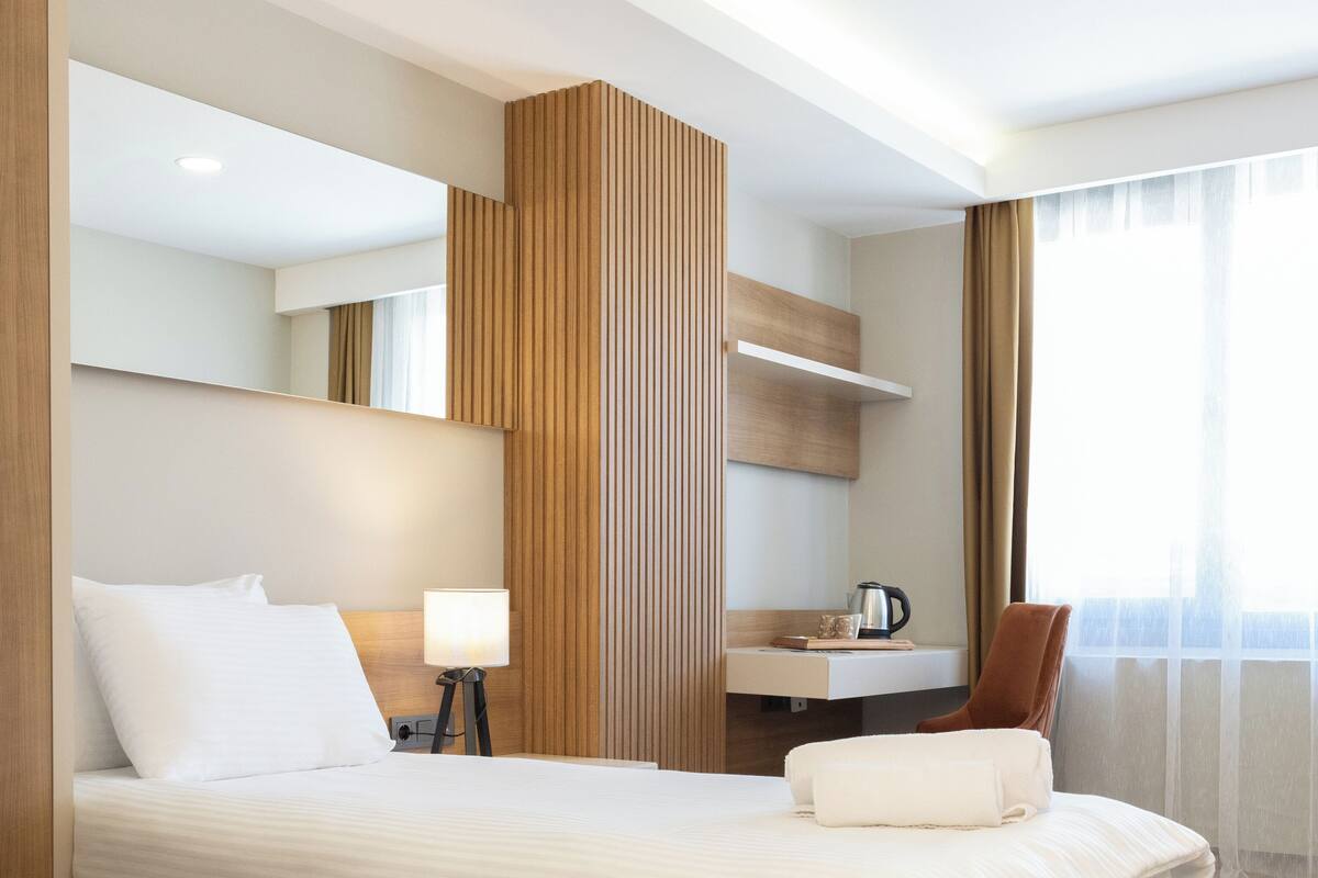 Une chambre d’hôtel moderne avec un grand lit, une table de chevet avec une lampe, un mur décoratif en bois, un petit coin salon et une vue sur l’extérieur.