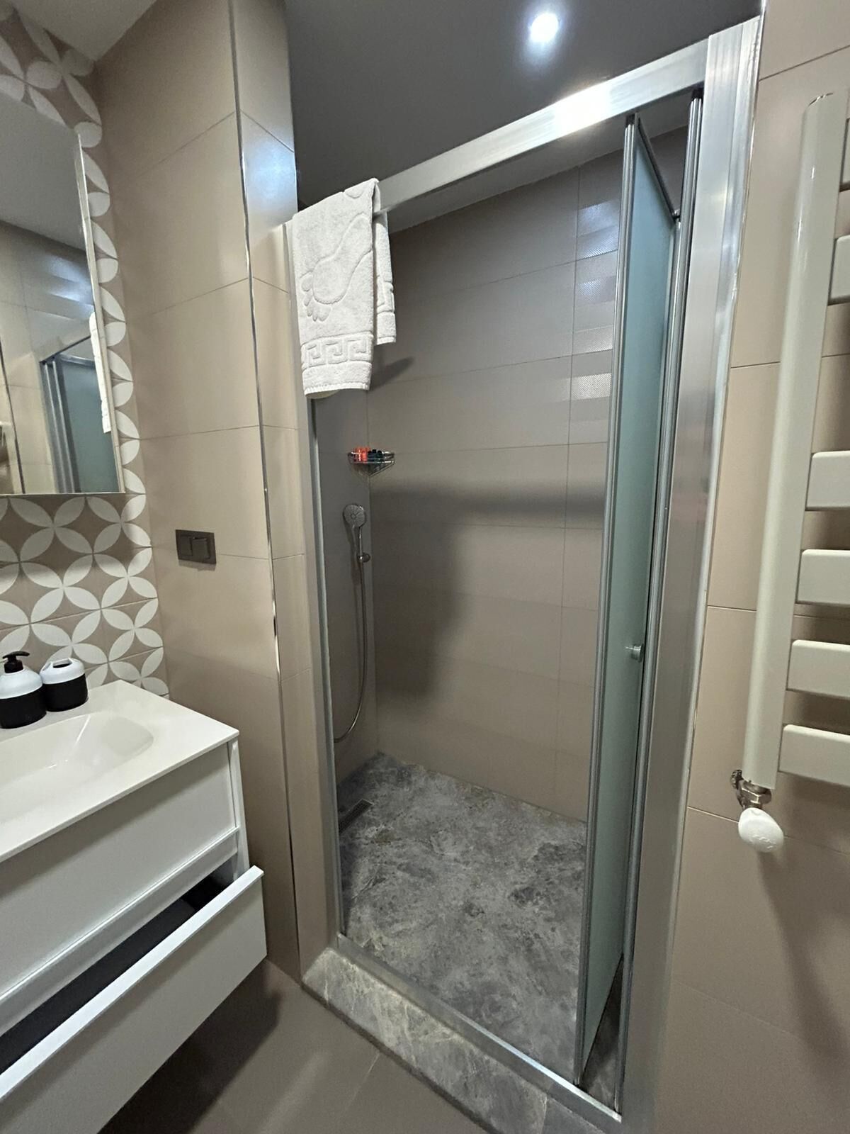 Un baño moderno con ducha de vidrio, lavabo blanco con dispensadores de jabón y un diseño de pared estampado.