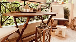 Un balcon avec une table en bois et deux tabourets, entouré de verdure et agrémenté d’une armoire blanche.