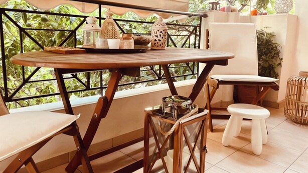 A balcony with a wooden table and two stools, surrounded by greenery and a white cabinet. - Modern Lux Apt by Acropolis I Private Parking, Gym, Balcony, Metro 1-min Walk (Athina)