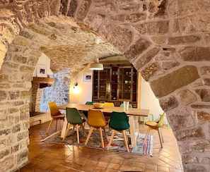 Uma zona de refeições rústica com um arco de pedra, mesa de madeira e cadeiras coloridas.