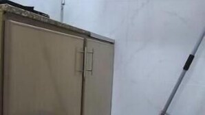 Un baño pequeño con azulejos, un armario beige, una escoba apoyada en la pared y un cubo de basura azul.
