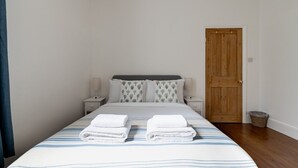 A neatly made bed with striped bedding and two towels, a wooden door, and a small bedside table with a lamp. - GuestReady - 2bed Battersea flat with Patio (London)