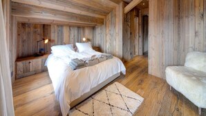 A bedroom with a wooden ceiling and walls, a bed with white linens, a fluffy white chair, and a small bedside table with a lamp. - 220sqm duplex luxury in the heart of Verbier (Val de Bagnes)