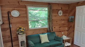 A cozy room with a green sofa, a small side table with a vase of flowers, a window with curtains, a clock on the wall, and a door. - Newly Renovated Cabin on a Private Lake Great for a Romantic Getaway or some R&R (Second Eel River Lake)