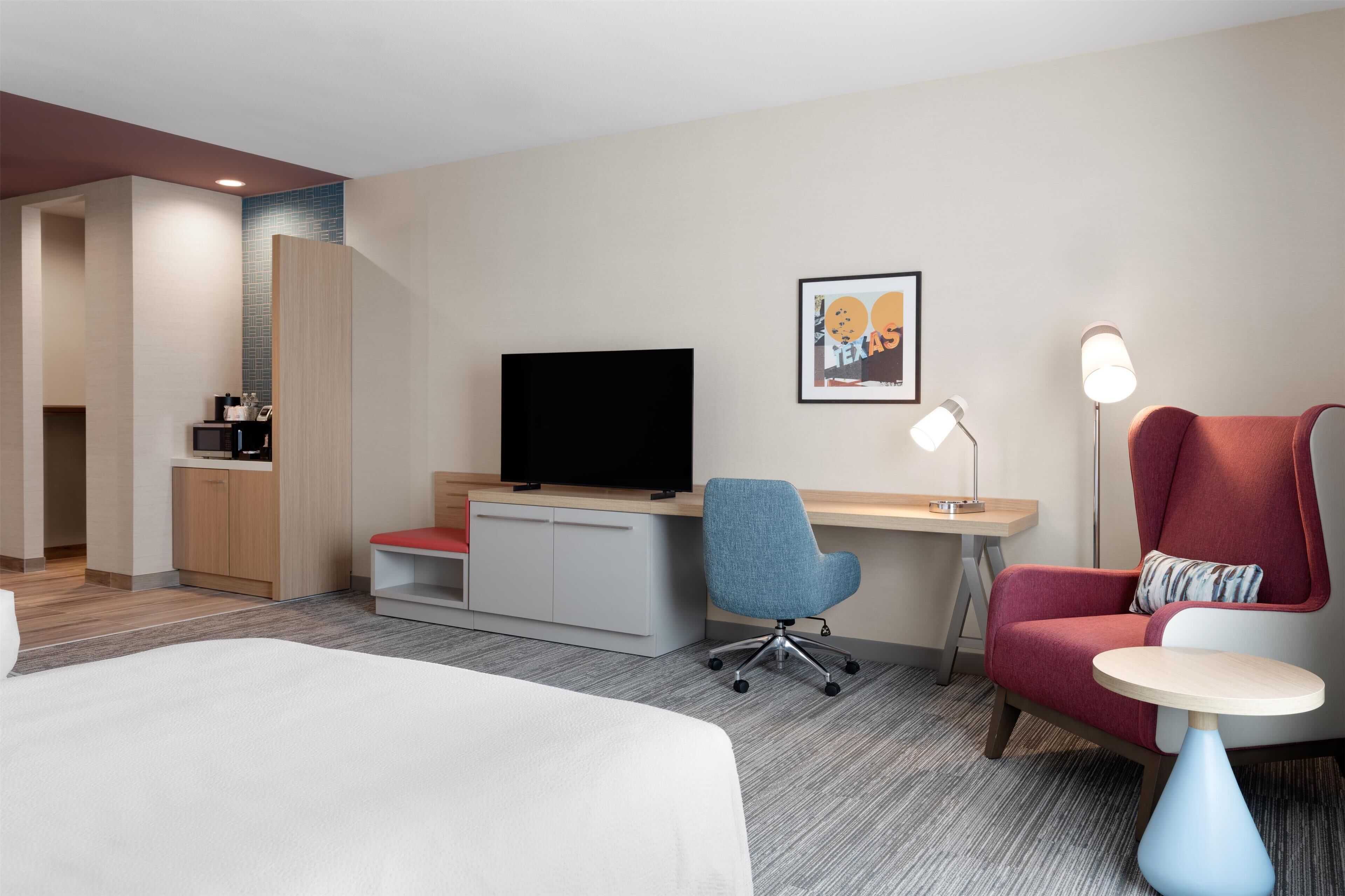 A hotel room with a bed, a desk, a chair, a TV, a small table, and a lamp.