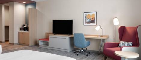 A hotel room with a bed, a desk, a chair, a TV, a small table, and a lamp.