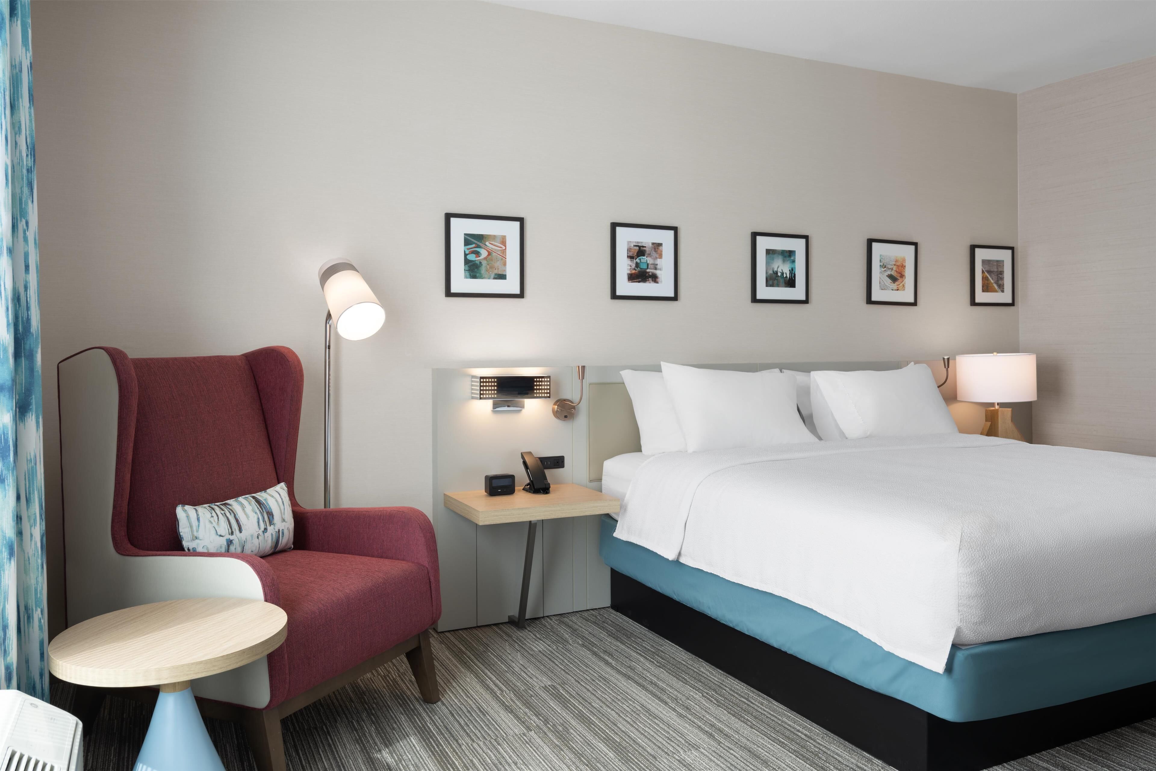 Ett hotellrum med en säng, ett skrivbord, en stol, en lampa och inramade tavlor på väggen.