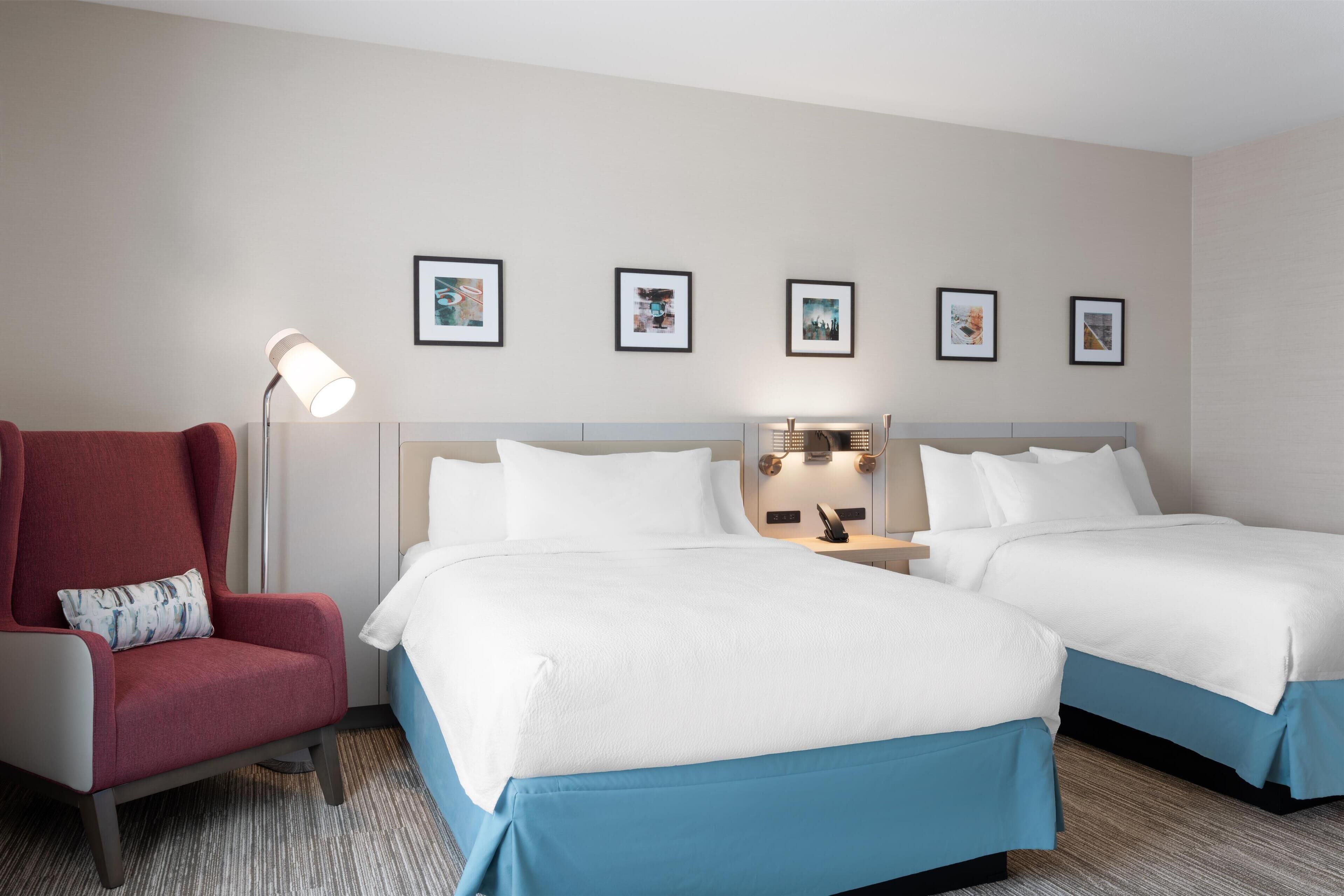 Habitación de hotel con dos camas, un sillón rojo, una lámpara de pie y cuatro cuadros en la pared.