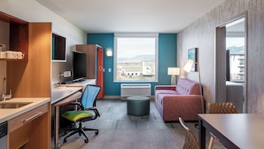A hotel room with a kitchenette, a TV, a sofa, a desk, and a large window offering a city view. - Home2 Suites by Hilton Bozeman Midtown (Bozeman)