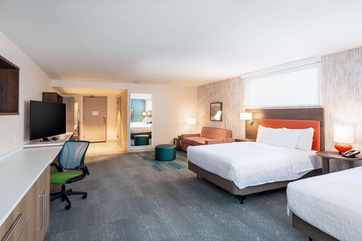 A hotel room with a desk, two beds, a TV, and a chair.