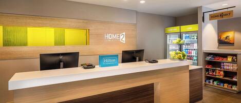 A hotel reception desk with a sign reading "HOME 2 SUITES BY MILTON" and a "Hilton HONORS" sign. There are two black chairs, a cash register, and a refrigerator with a "really cold" sign.