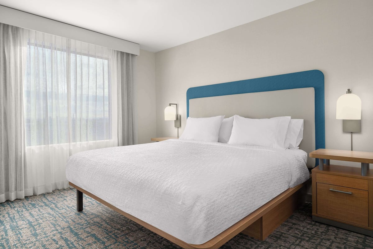 A hotel room with a large bed, a wooden nightstand, a lamp, and a view of the outdoors through sheer curtains.