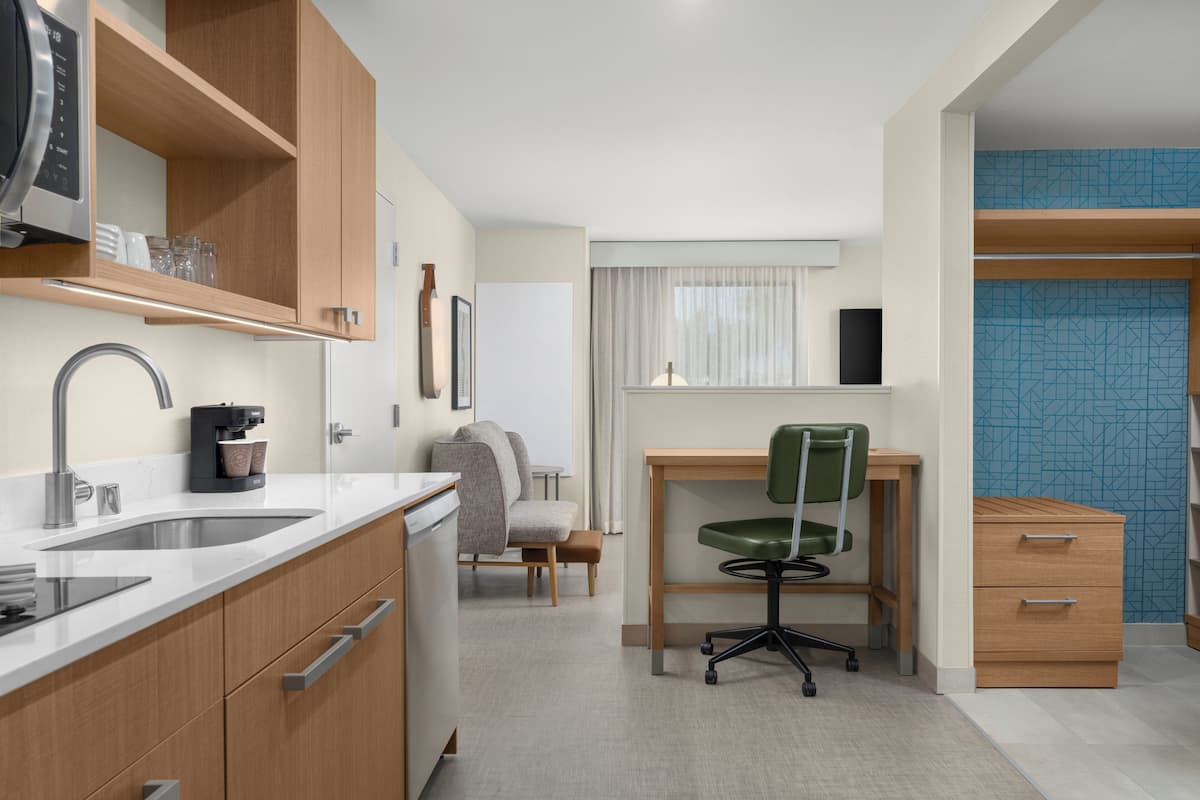 A modern kitchen with a built-in coffee machine, a sink, and wooden cabinets. There is a dining area with a table and chairs, and a small seating area with a chair and a sofa.