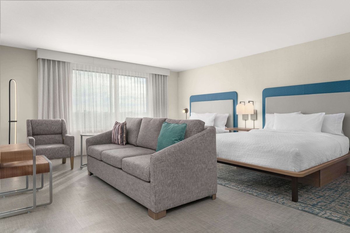 A hotel room with a grey sofa, a wooden chair, a bed with a blue headboard, and a nightstand with lamps.