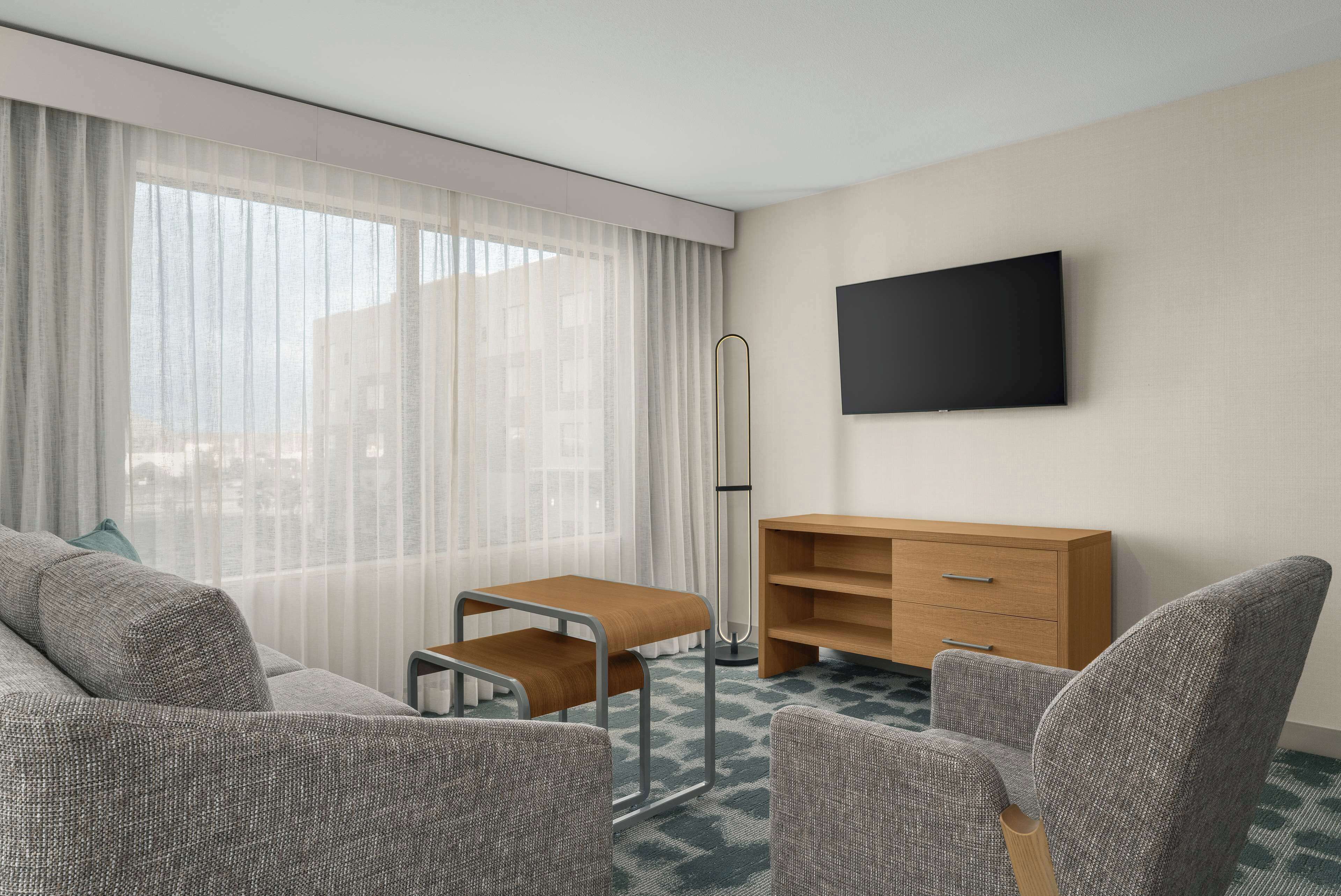 A hotel room with a sofa, armchairs, a small wooden table, a TV mounted on the wall, and a floor lamp.
