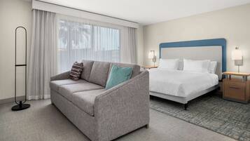 A hotel room with a grey sofa, a bed with a blue headboard, a bedside table with a lamp, and a floor lamp.